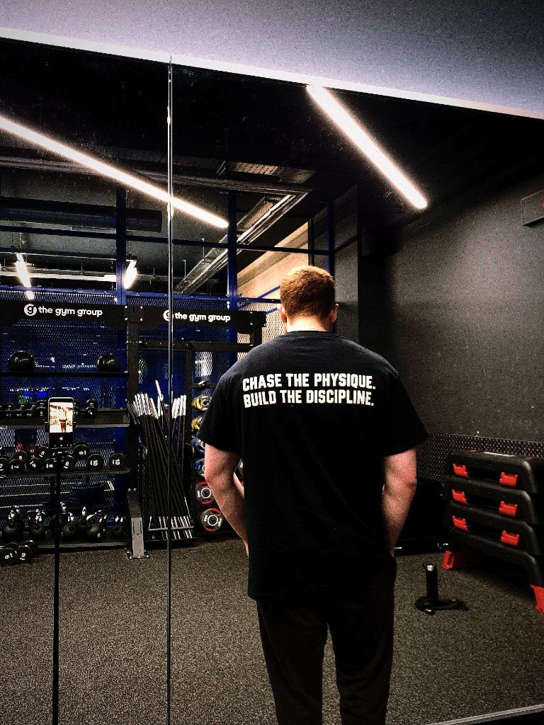 Davide from behind at The Gym Group White City. His shirt reads Chase the Physique. Build the Discipline.